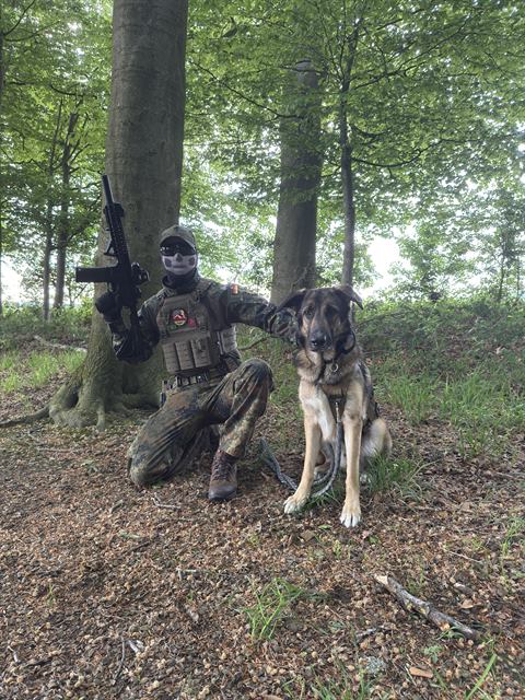 OMUNE kniet in Flecktarn-Ausrüstung mit Replik neben einem Schäferhund im Wald