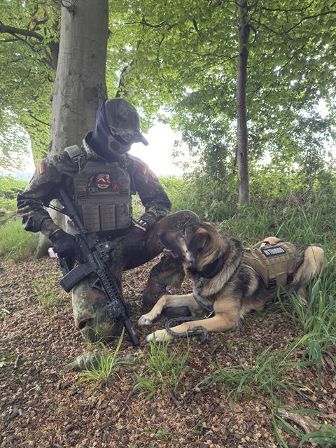 OMUNE kniet mit Replik im Wald, der Hund mit Hundeweste liegt daneben